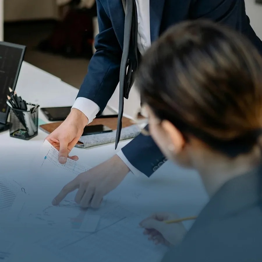 A male employee pointing towards a graph on paper with a female employee watching and taking notes.
